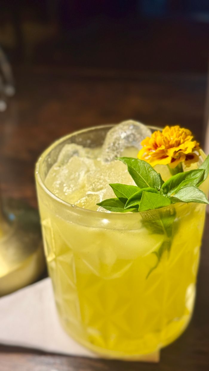 Contact Close-up of a yellow cocktail with ice, basil leaves, and a marigold garnish on a dark wooden table.